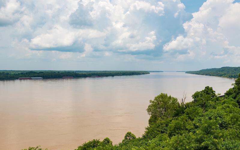 Wide, muddy river with barges, flanked by lush green trees under a cloudy sky.
