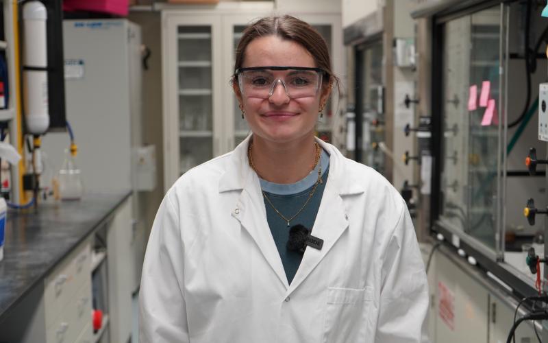 Young woman in lab coat and safety glasses smiles in a laboratory setting.