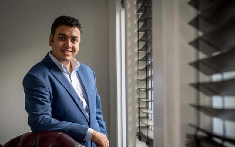 Michael Naguib in a blue suit leans on a leather chair next to a window.