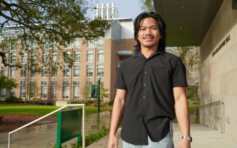 Young man in a black shirt smiles on a campus.