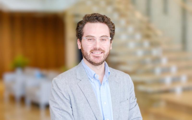 A smiling man in a grey suit jacket and light blue shirt stands in front of a blurred staircase.