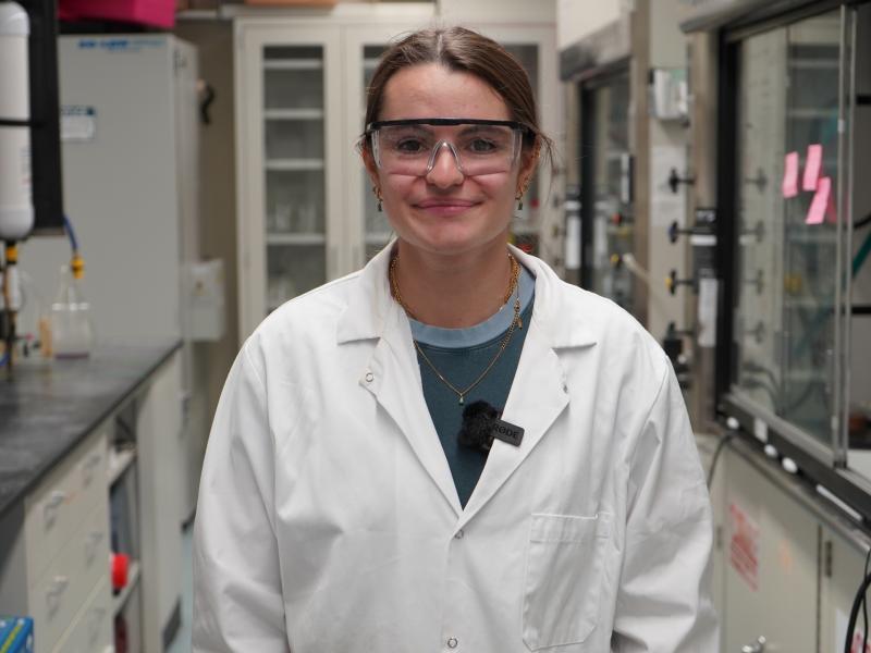 Young woman in lab coat and safety glasses smiles in a laboratory setting.