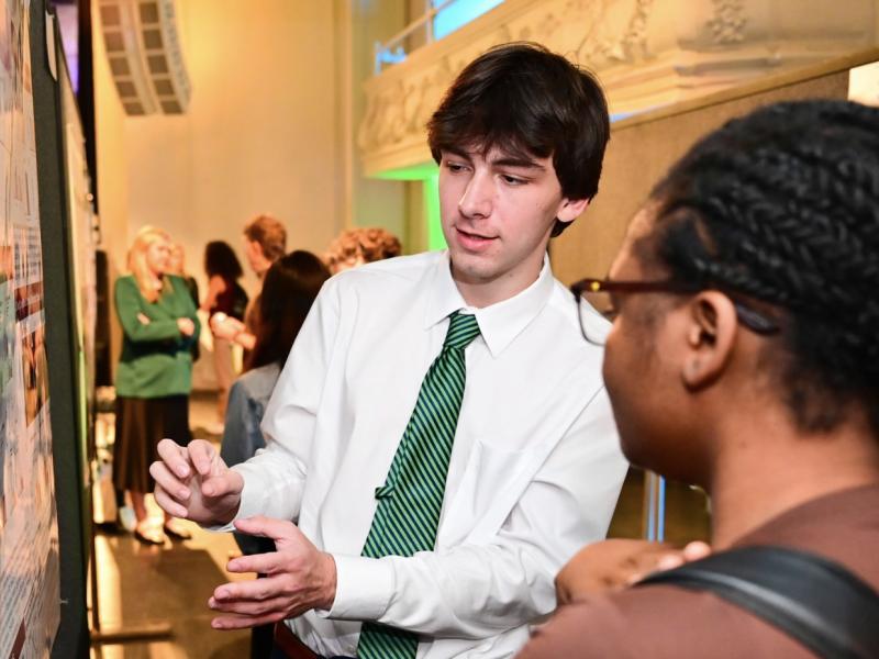 Man in a white shirt and green tie explains a poster to a woman in glasses.