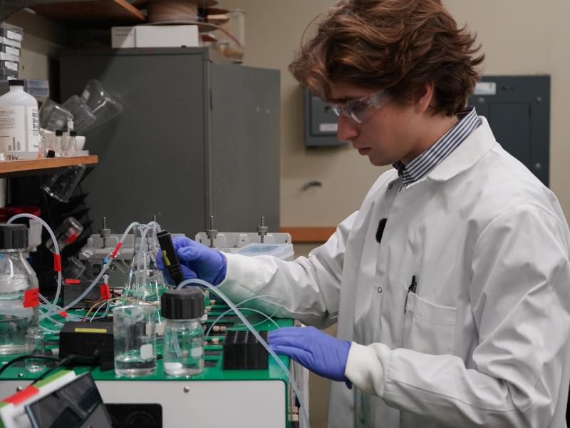 A male scientist in a lab coat and goggles works with scientific equipment.