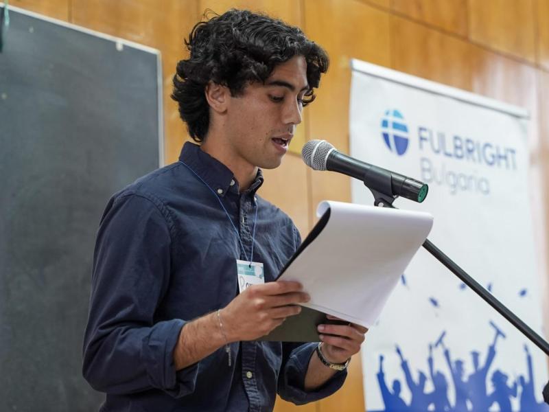 Young man speaks into a microphone, holding a paper. Fulbright Bulgaria banner in background.