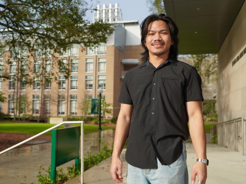 Young man in a black shirt smiles on a campus.