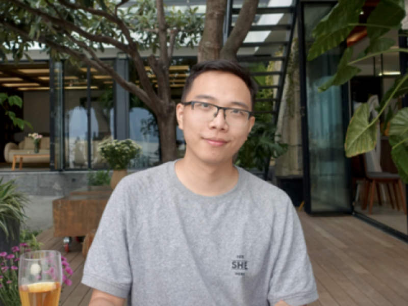 Young man in a grey t-shirt and glasses smiles at the camera, outdoors.