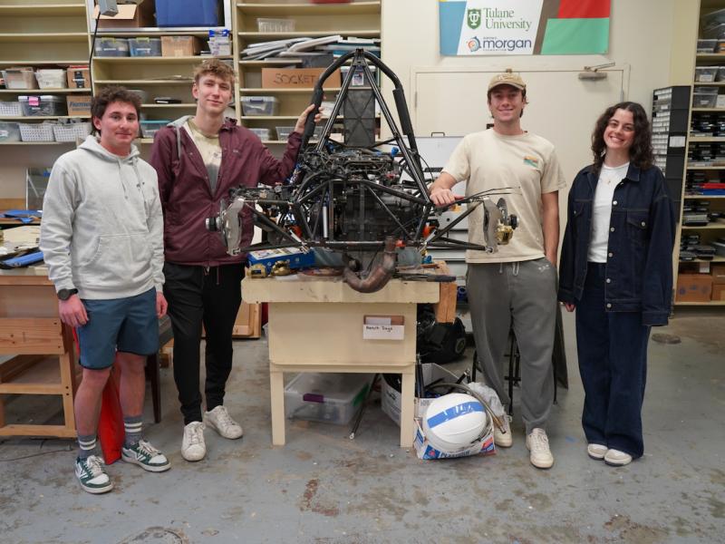 Four smiling students stand around a large, complex machine in a workshop.