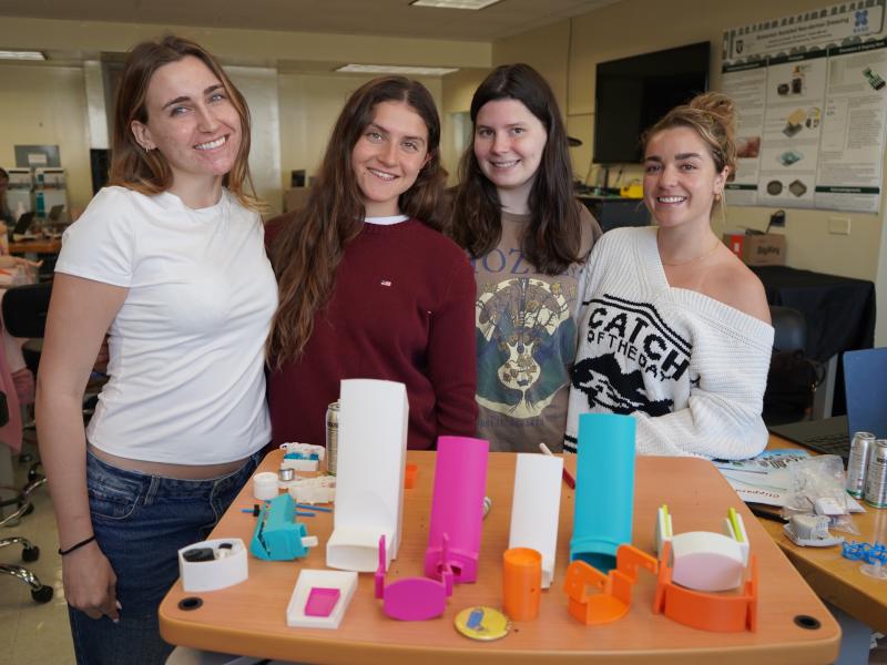 Four smiling women stand behind a table displaying colorful 3D-printed objects.