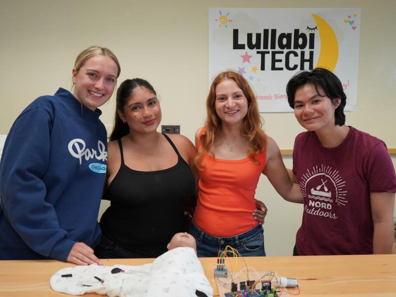 Four smiling young women pose for a photo behind a table with a baby doll and tech equipment.