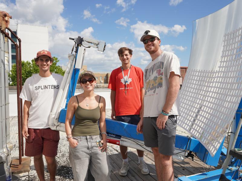 Four smiling people in casual clothing stand on a rooftop with a large solar panel.