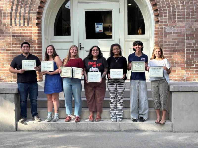 Seven students, holding certificates, stand on steps in front of a building.