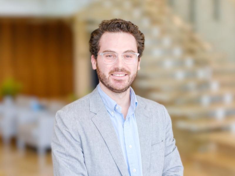A smiling man in a grey suit jacket and light blue shirt stands in front of a blurred staircase.