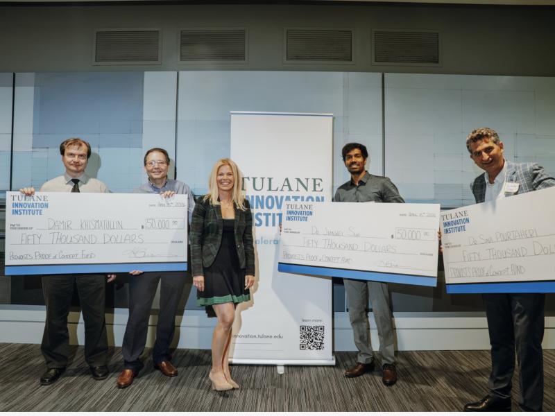 Five people smile while holding large checks in front of a banner.