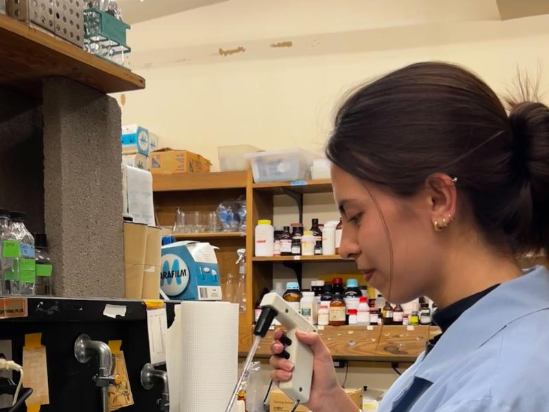 A woman in a lab coat uses a pipette to transfer liquid into a flask.