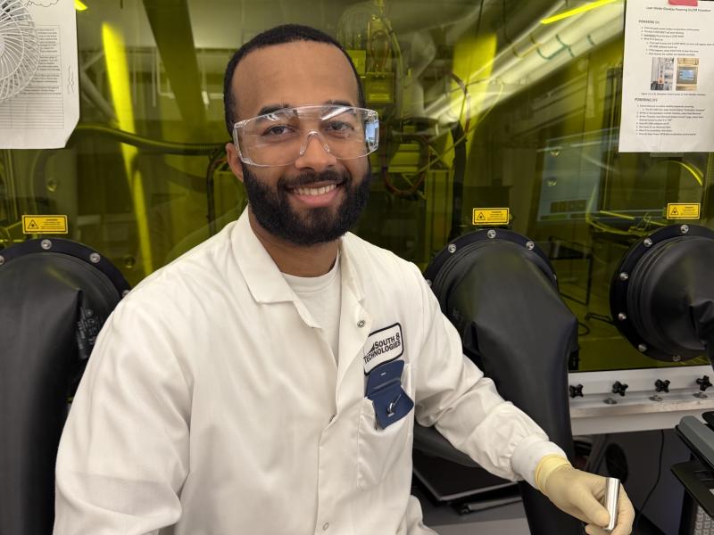 Man in lab coat and goggles smiles, wearing gloves and holding a tool.