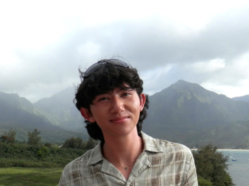 Young man in patterned shirt smiles on a balcony with green mountains and ocean background.