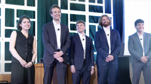 Five smiling people on a stage, dressed in suits.