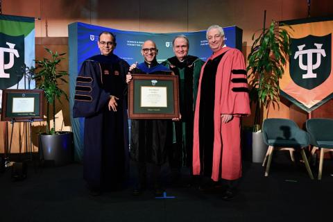 Four men, three in academic robes, present a framed award at a university ceremony.