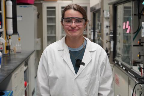 Young woman in lab coat and safety glasses smiles in a laboratory setting.