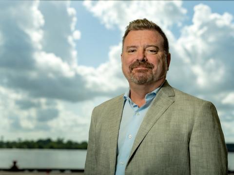 Photograph of a man in a suit standing in front of a cloudy sky.