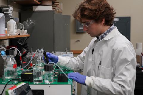 A male scientist in a lab coat and goggles works with scientific equipment.