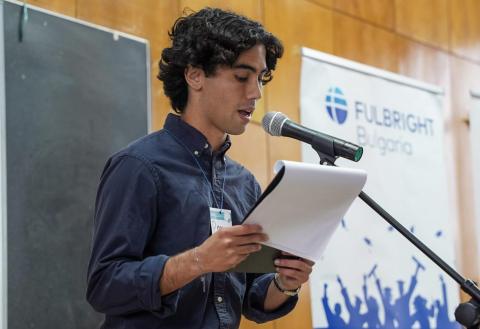 Young man speaks into a microphone, holding a paper. Fulbright Bulgaria banner in background.