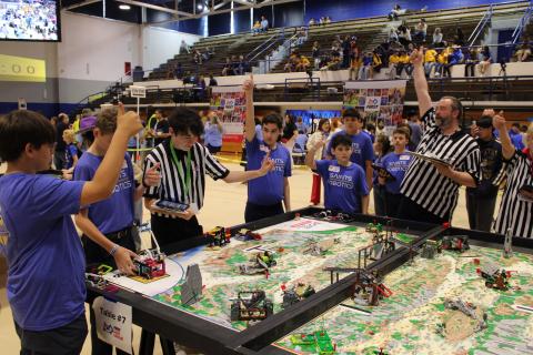 Students and referees celebrate near a detailed robot competition table.