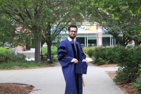 Graduate in blue gown and cap stands on a pathway between trees.