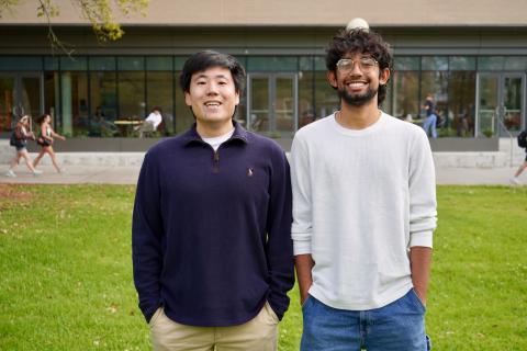 Two smiling young men stand on a grassy lawn in front of a building.