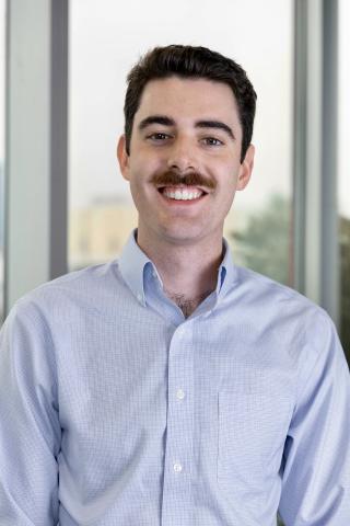 A young man with a mustache wears a light blue button-down shirt and smiles at the camera.