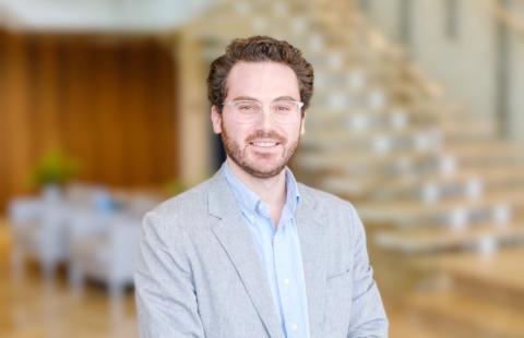 A smiling man in a grey suit jacket and light blue shirt stands in front of a blurred staircase.