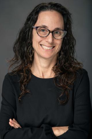 Smiling woman with dark curly hair and glasses, arms crossed.