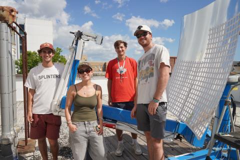 Four people stand on a rooftop near a large white satellite dish on a blue lift.
