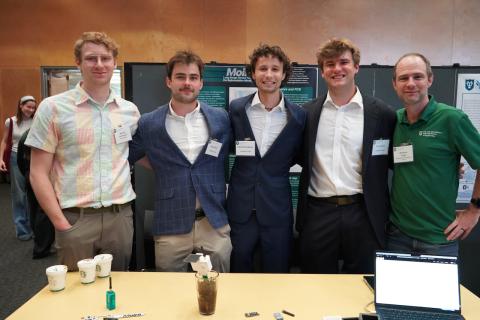 Five young men pose at a table with a laptop and drinks.