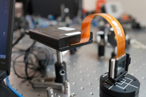 Close-up of a flexible circuit board connected to a small black device on a lab bench.