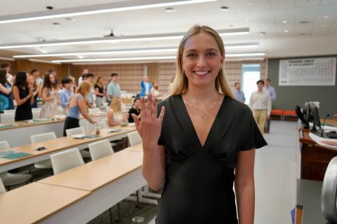 Young woman in black dress waves to the camera in a classroom with other students.