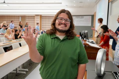 Man in green shirt waves hello in classroom with audience.