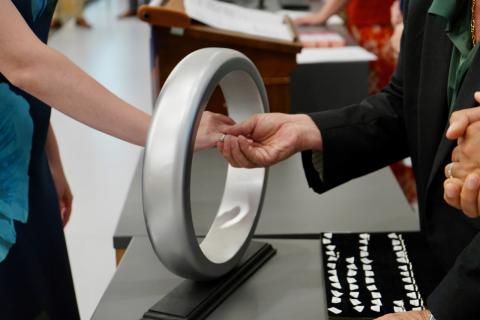 Hands exchanging a ring through a modern, silver ring sculpture.
