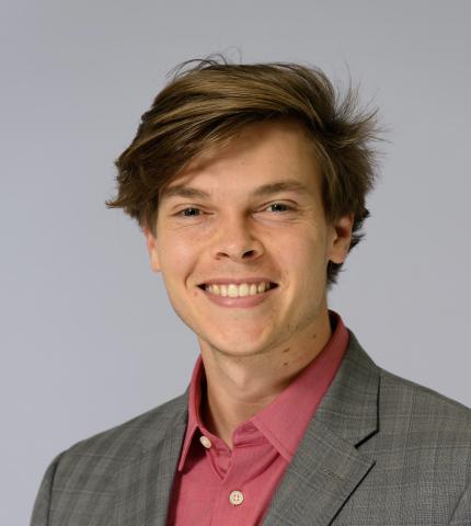 A young man in a gray suit and pink shirt smiles at the camera.