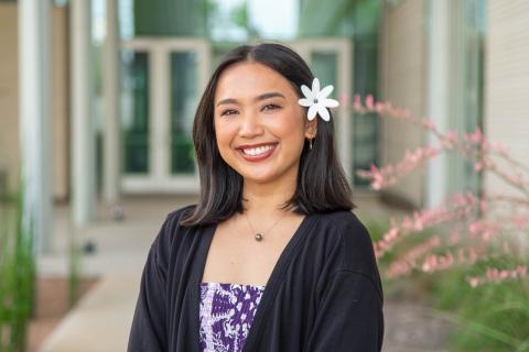 Young woman with a white flower in her dark hair, smiling, wearing a purple and white patterned top and a black cardigan.