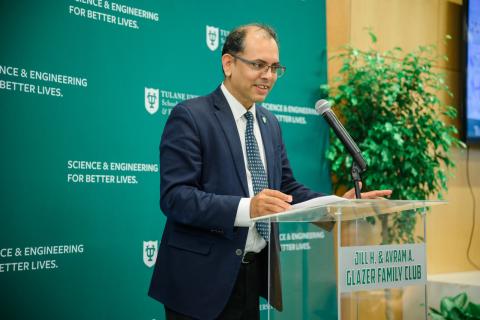 Man in a suit speaks at a podium with a green background and plants.