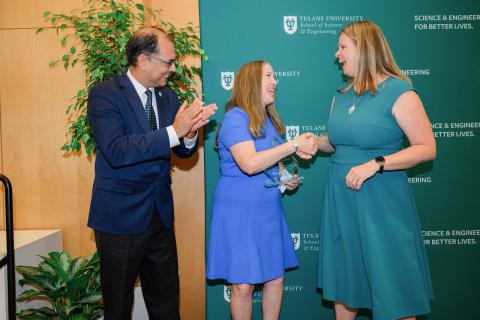 Woman in blue dress accepts award from woman in teal dress, with man in suit clapping.