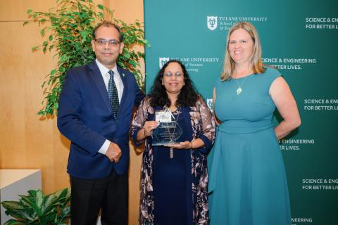 Three people stand together. The person in the middle holds an award.