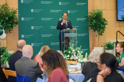 Man in suit speaks at podium with green backdrop.