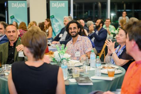 A smiling man with curly hair at a dinner event, with people in the background and Dasani logos.