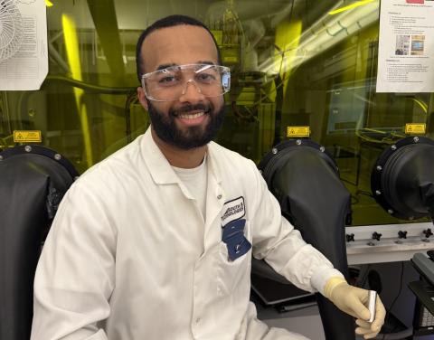 Man in lab coat and goggles smiles, wearing gloves and holding a tool.