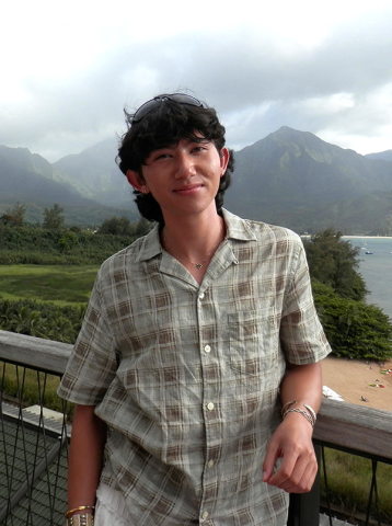 Young man in patterned shirt smiles on a balcony with green mountains and ocean background.
