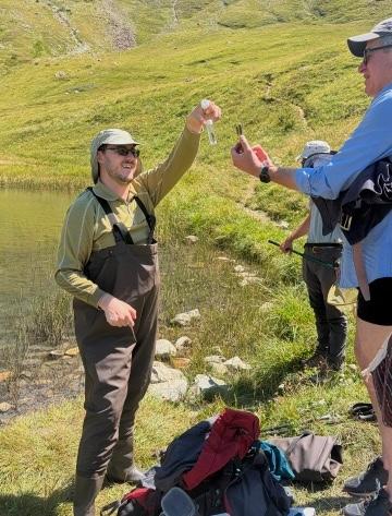 Two people in waders examine a small vial in a grassy, mountainous area near a lake.