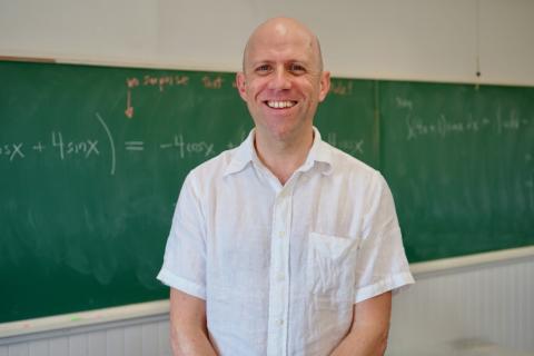 Photograph of a smiling bald man in a white shirt standing before a green chalkboard.